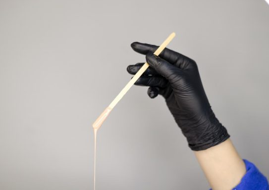 Black gloved hand holding a wooden stick with a viscous substance dripping from it.